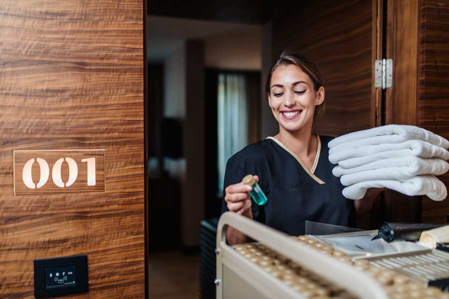 hotel maid in uniform bringing clean towels and other supplies to hotel room.